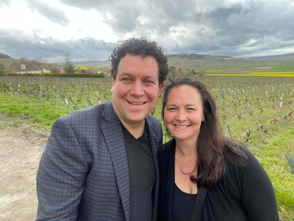 Marcus and Jamie Guiliano smile together while standing in a vineyard, with rolling hills and yellow fields visible in the background under a cloudy sky.