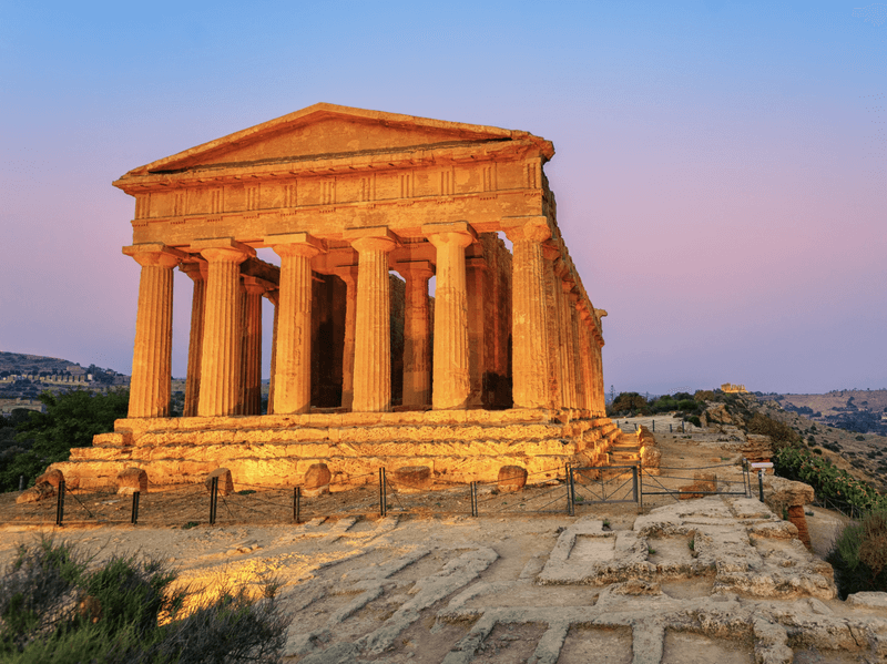 The well-preserved Temple of Concordia stands majestically against a soft pink and blue twilight sky, its golden stone pillars and pediment bathed in the warm light of dusk.