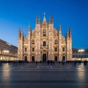 Duomo di Milano glowing at blue hour with subtle motion blur of passersby, elegant postcard-style travel photograph.