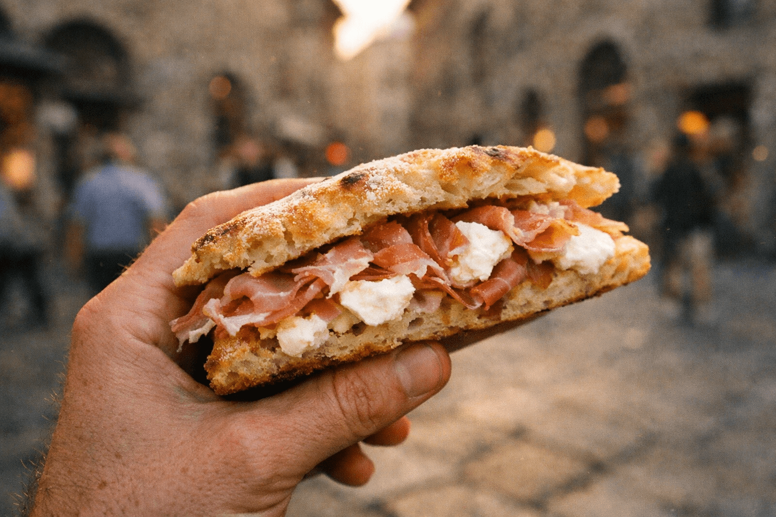 Hand holding a fresh schiacciata panino with prosciutto and mozzarella against a blurred Florence street background, representing authentic Italian street food culture.