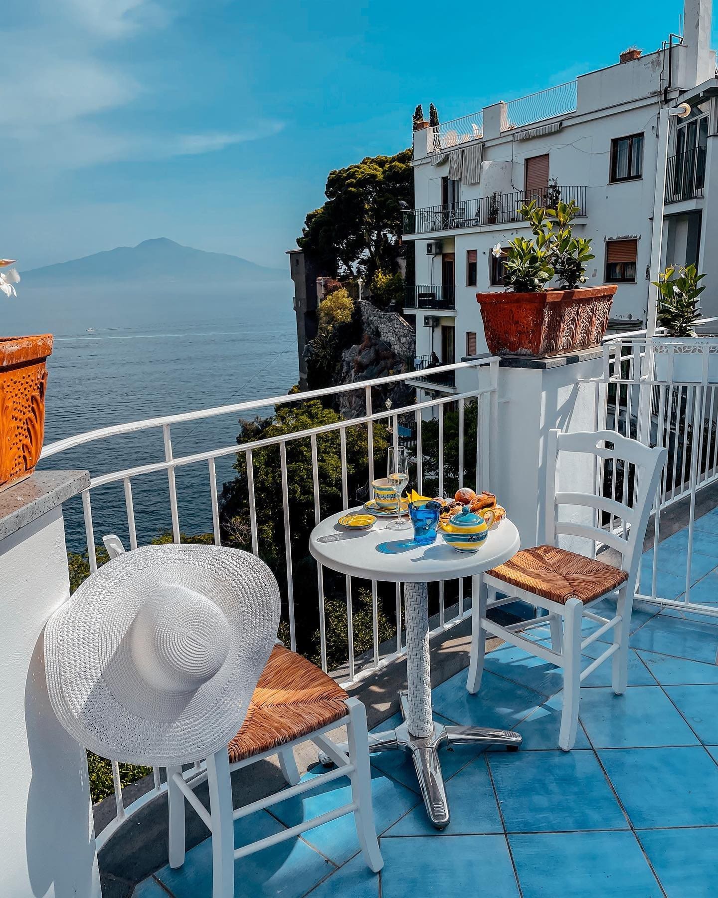 A balcony at Boutique Hotel Helios in Sorrento with a breakfast table, chairs, a white hat, overlooking the sea and a distant
