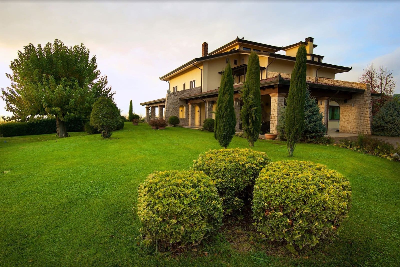 Radici Resort villa in Campania with a light facade, stone columns, green lawn, and tall cypress trees under a bright sky.