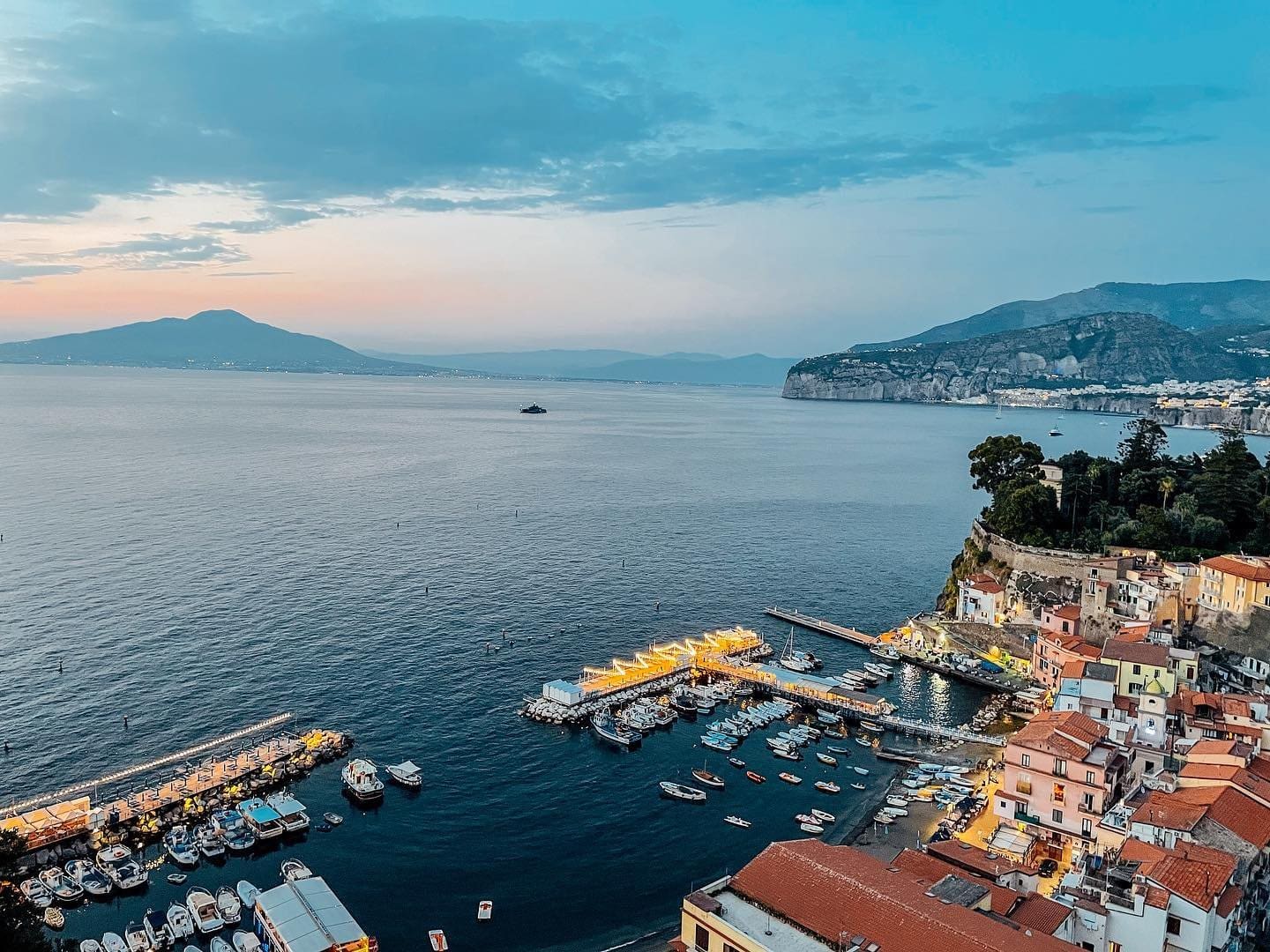 Elevated evening view of Sorrento, Campania, with an illuminated marina, colorful cliffside buildings, and Mount Vesuvius