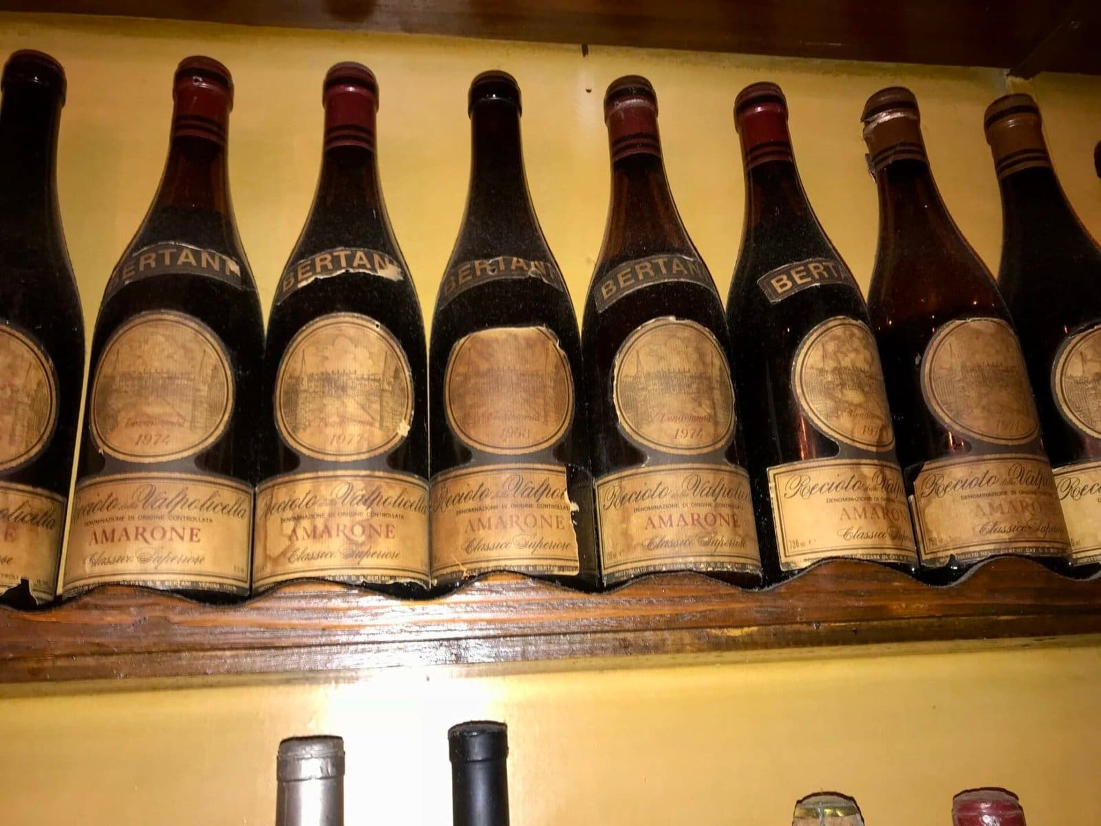Row of vintage Bertani Amarone wine bottles on a wooden shelf against a yellow wall, showing various years.