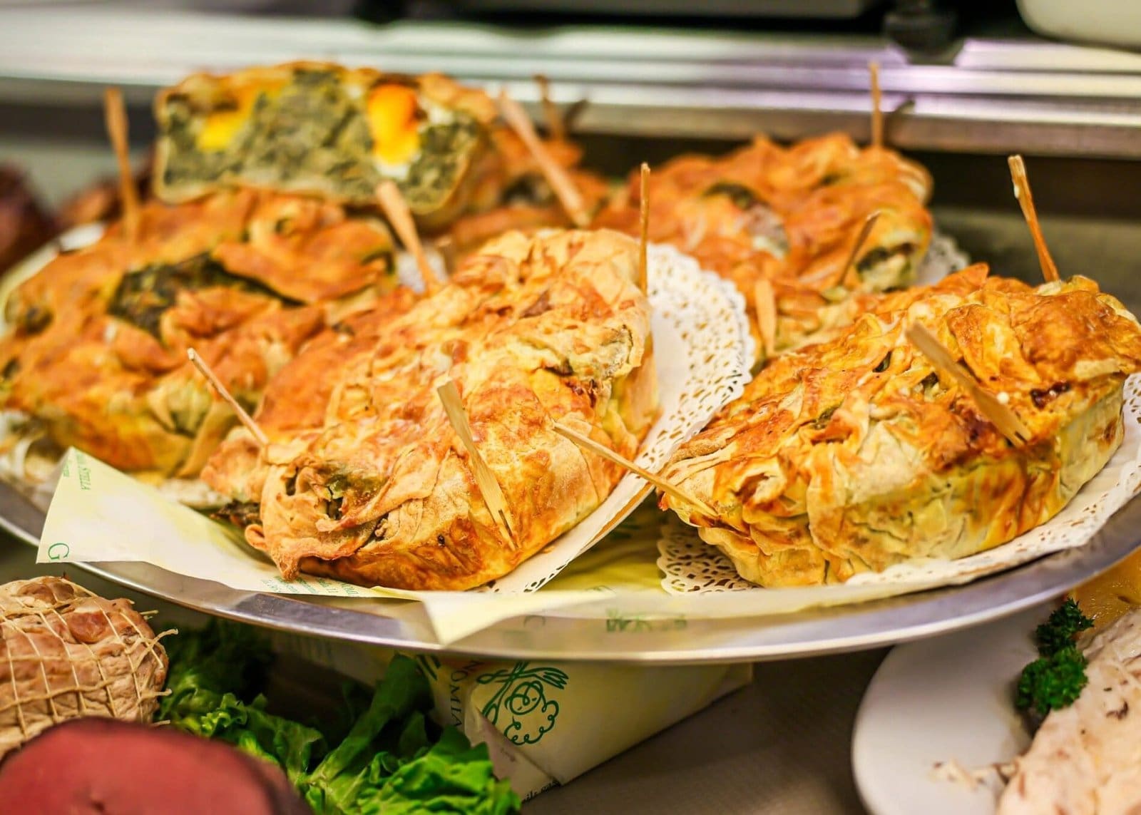 A deli tray of golden-brown savory pastries with green fillings, along with meats and fish, at Verona Deli.