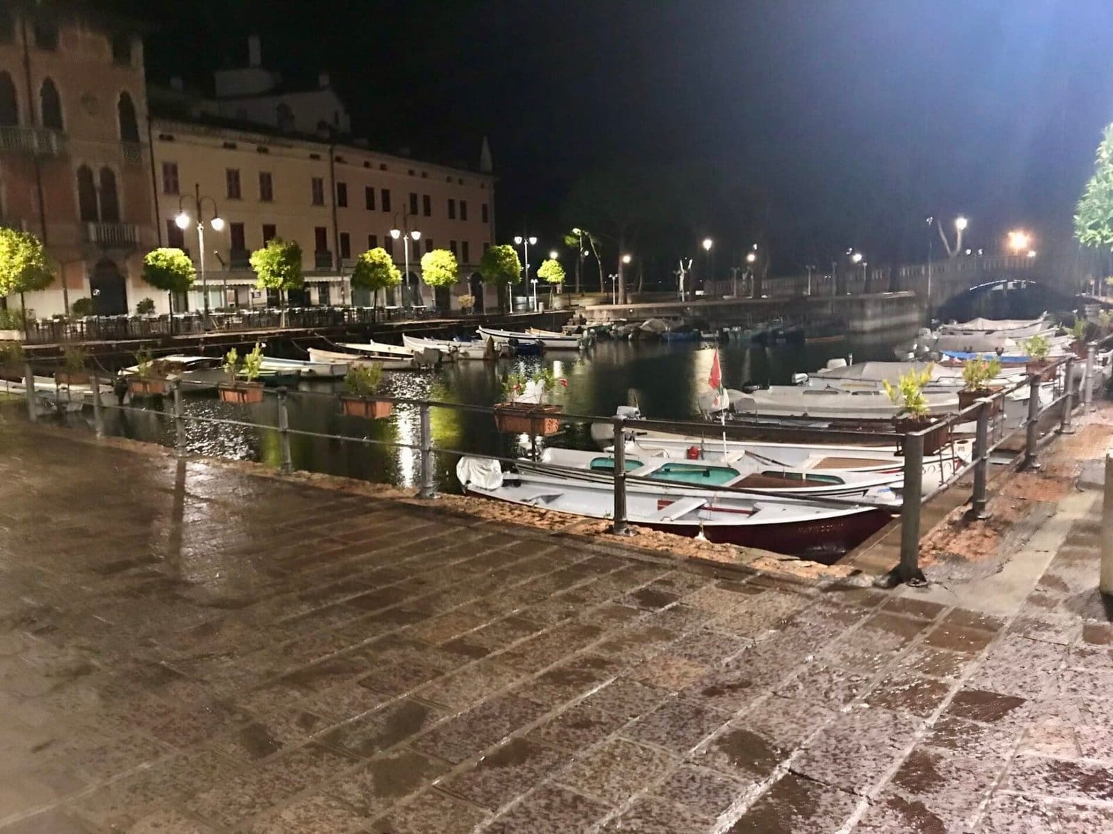 Night view of Lake Garda harbor with boats, illuminated buildings, and wet cobblestone pavement reflecting lights.