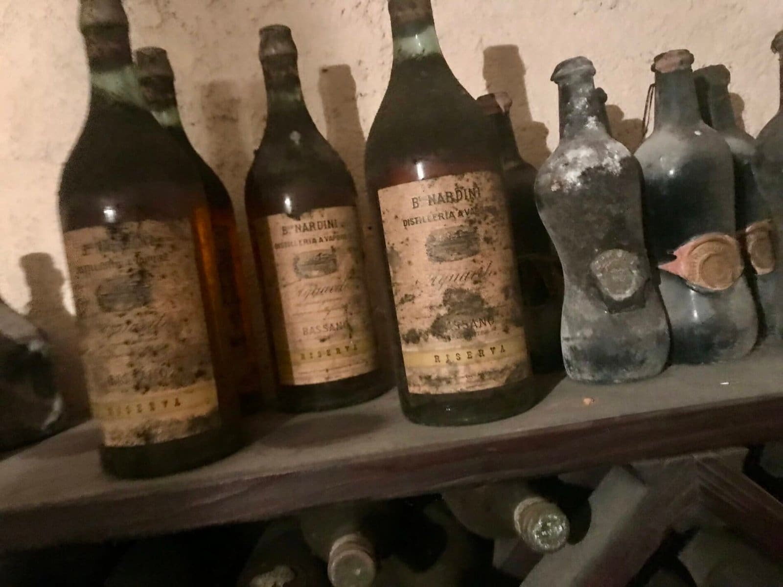 Dusty vintage Nardini Grappa Riserva bottles from Bassano on a wooden cellar shelf.