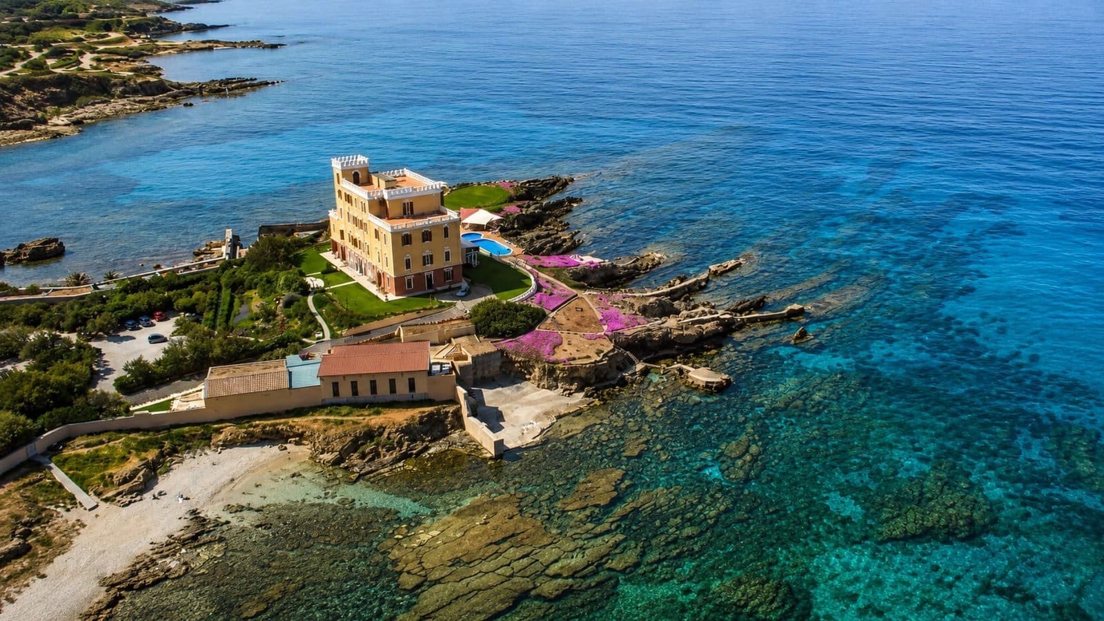 Aerial view of a yellow villa with a pool and pink flowers on a rocky coastline with clear turquoise water in Sardinia.