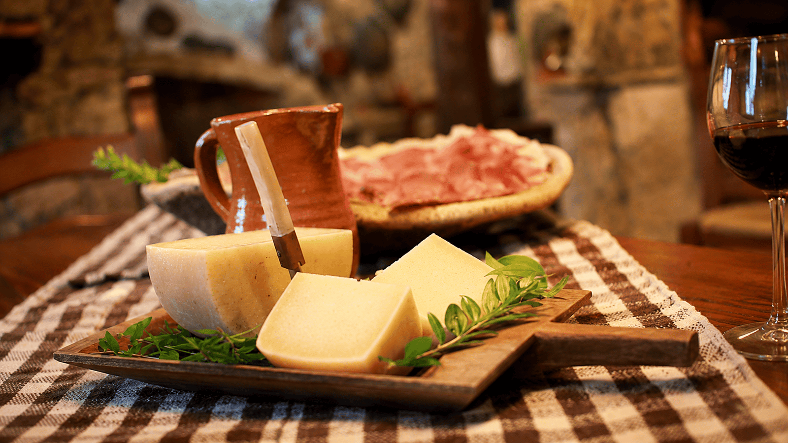 Sardinian cheese wedges and salumi platter with a terracotta pitcher and a glass of red wine on a checkered tablecloth.